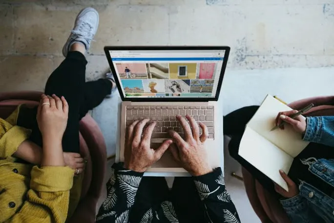 Two people reviewing content on a laptop while one takes notes in a notebook.