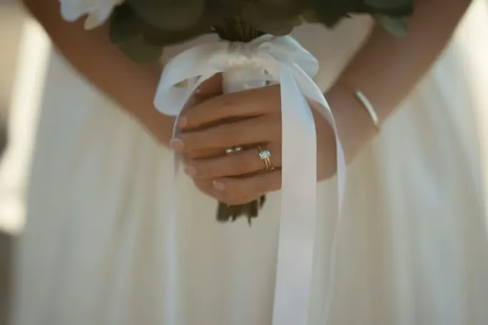 Bride holding a white floral bouquet, with a wedding ring visible on her hand.