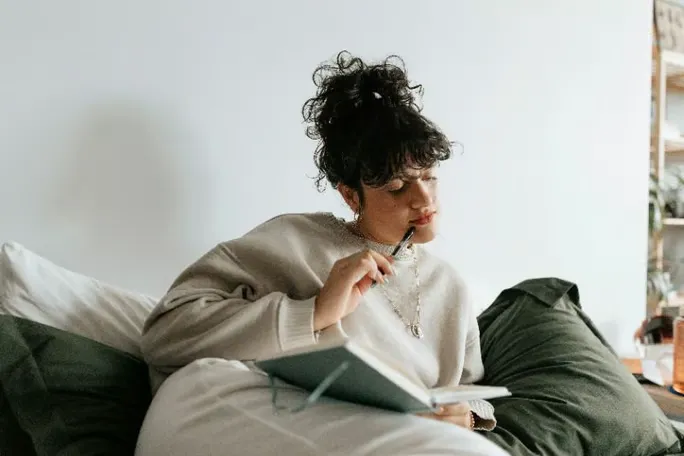 Woman sitting on a couch, thinking and writing in an open notebook.