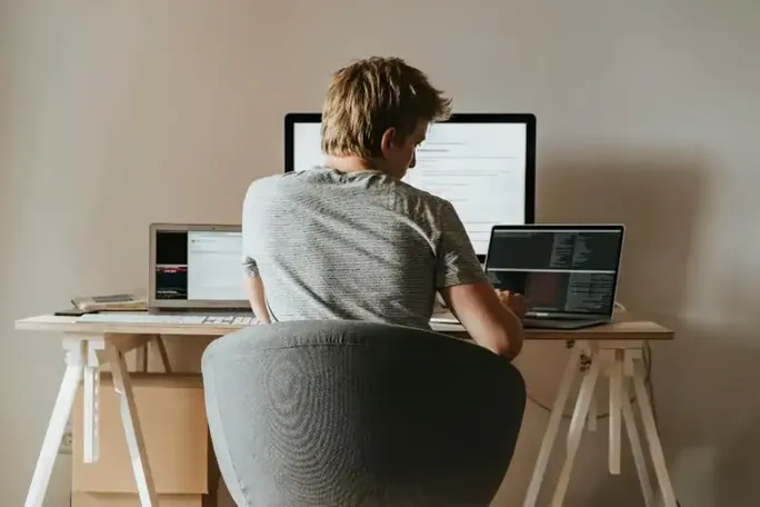 A man sitting at a wooden desk, simultaneously working on three computers.
