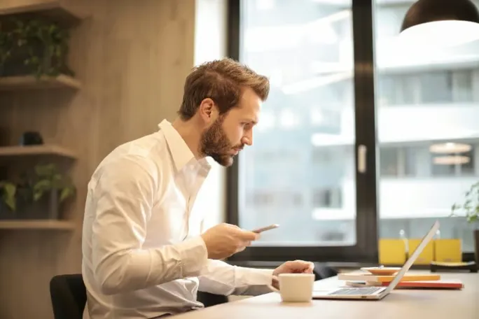 A man in a white shirt sitting at a desk working on a laptop and smartphone.