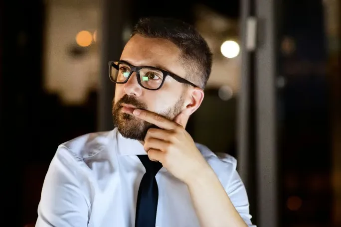 A man with glasses and a beard thinking with his hand on his face.