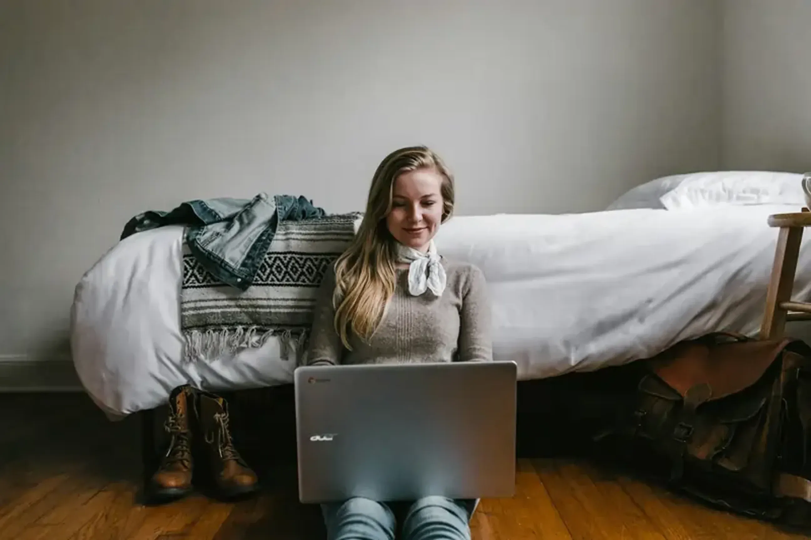 Woman sitting on the floor using a laptop by her bed.