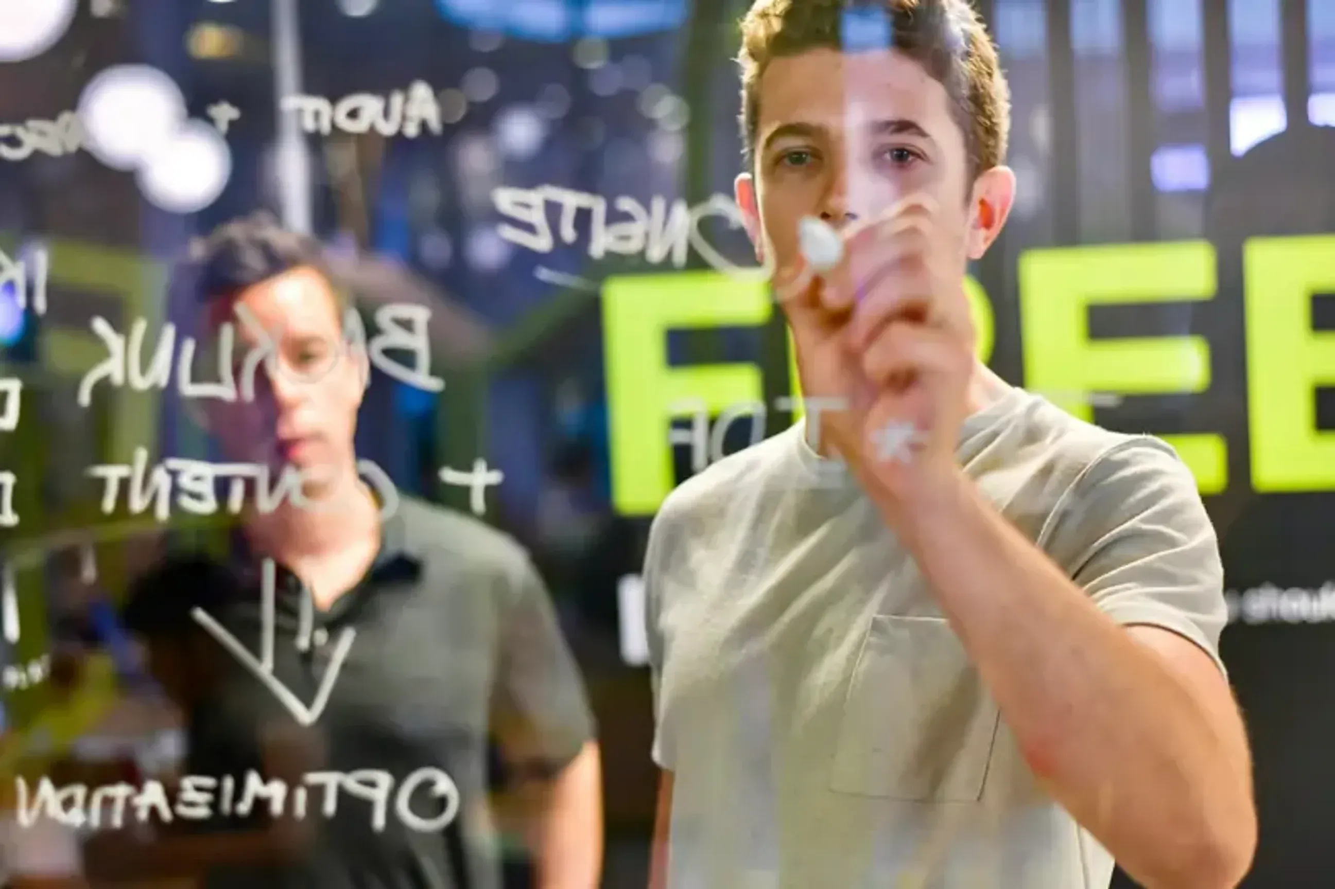 Man writing ideas on a glass board while a coworker watches.