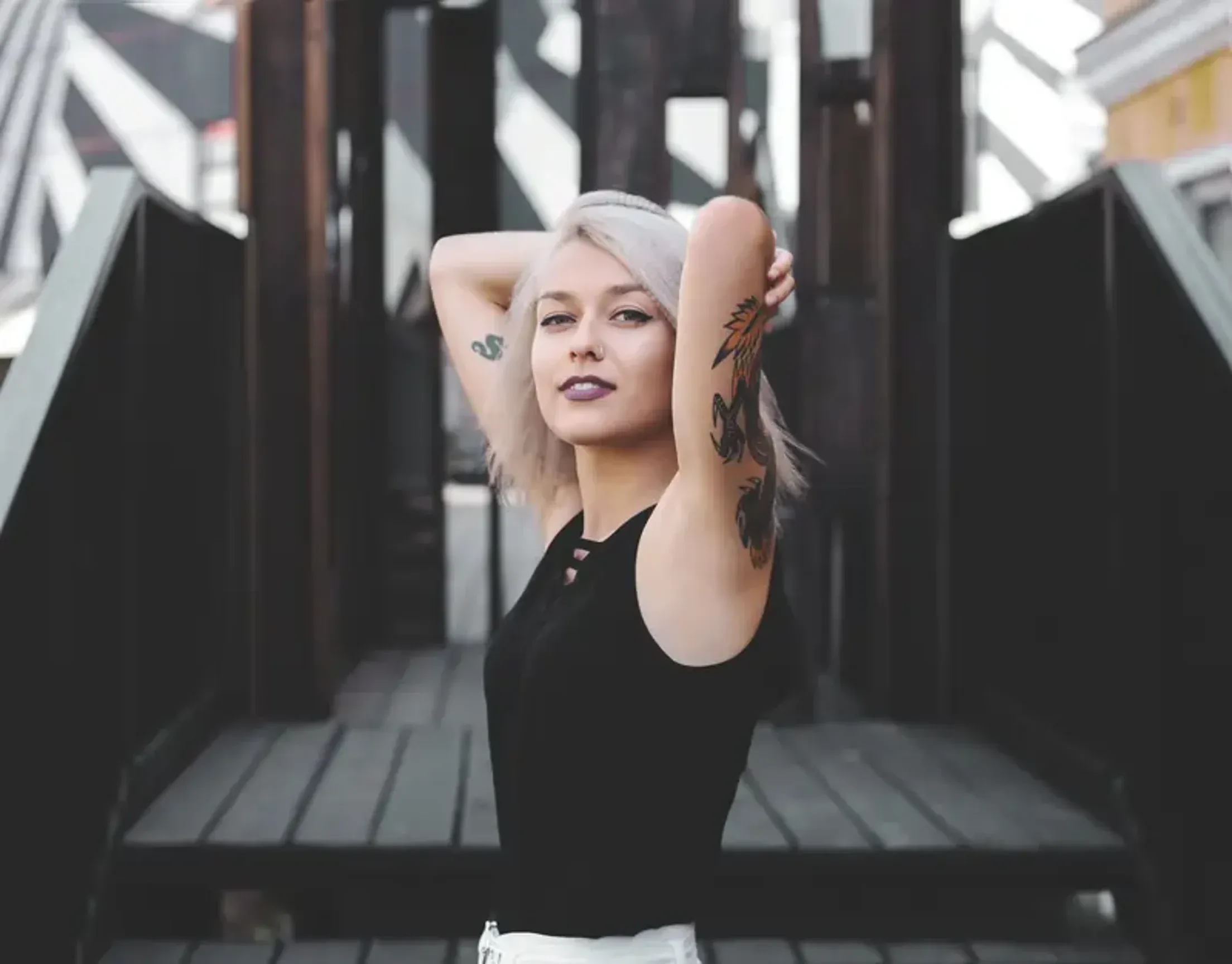 Woman with tattoos posing outdoors with hands behind her head.