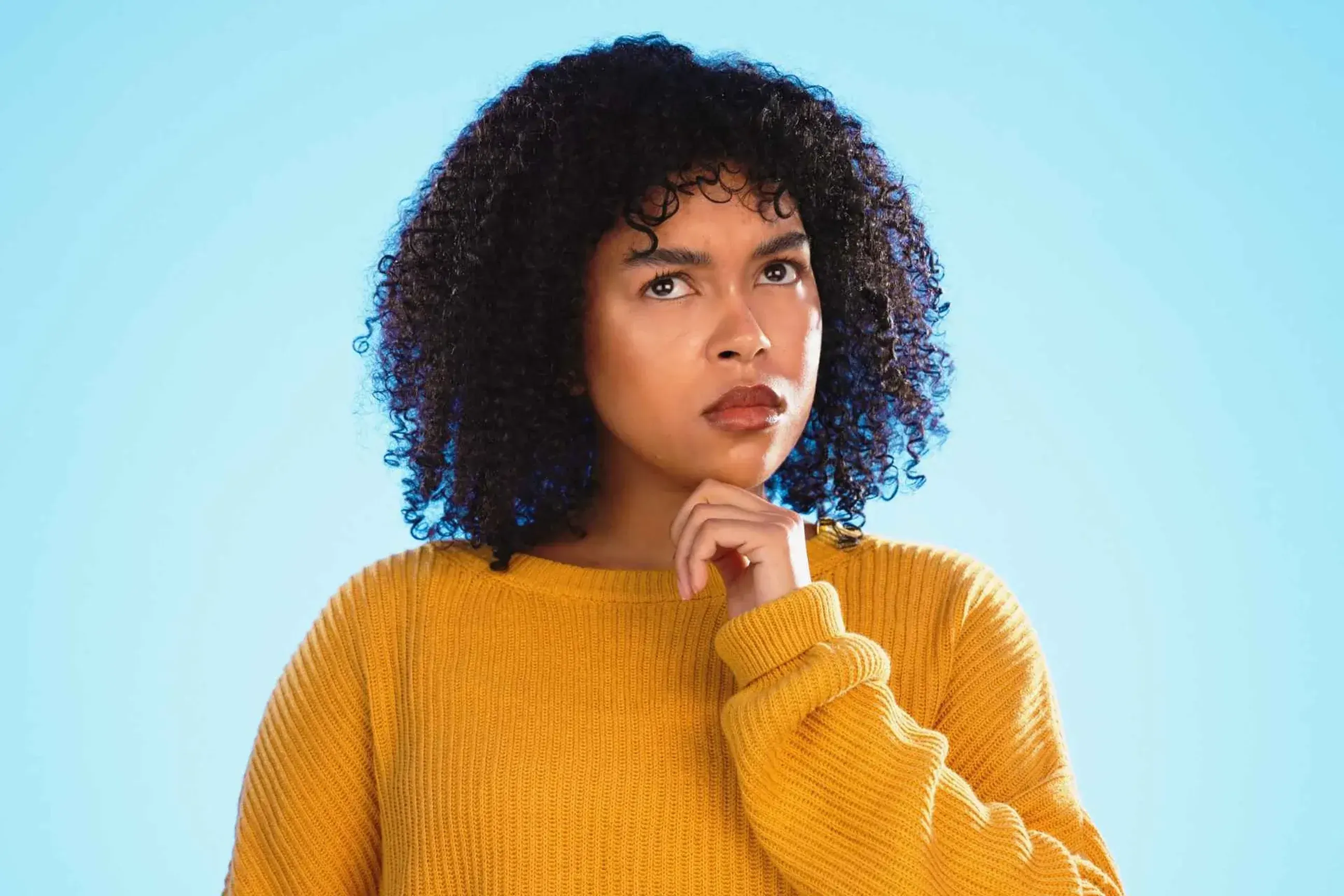 Thoughtful woman in a yellow sweater looking upward against a blue background.