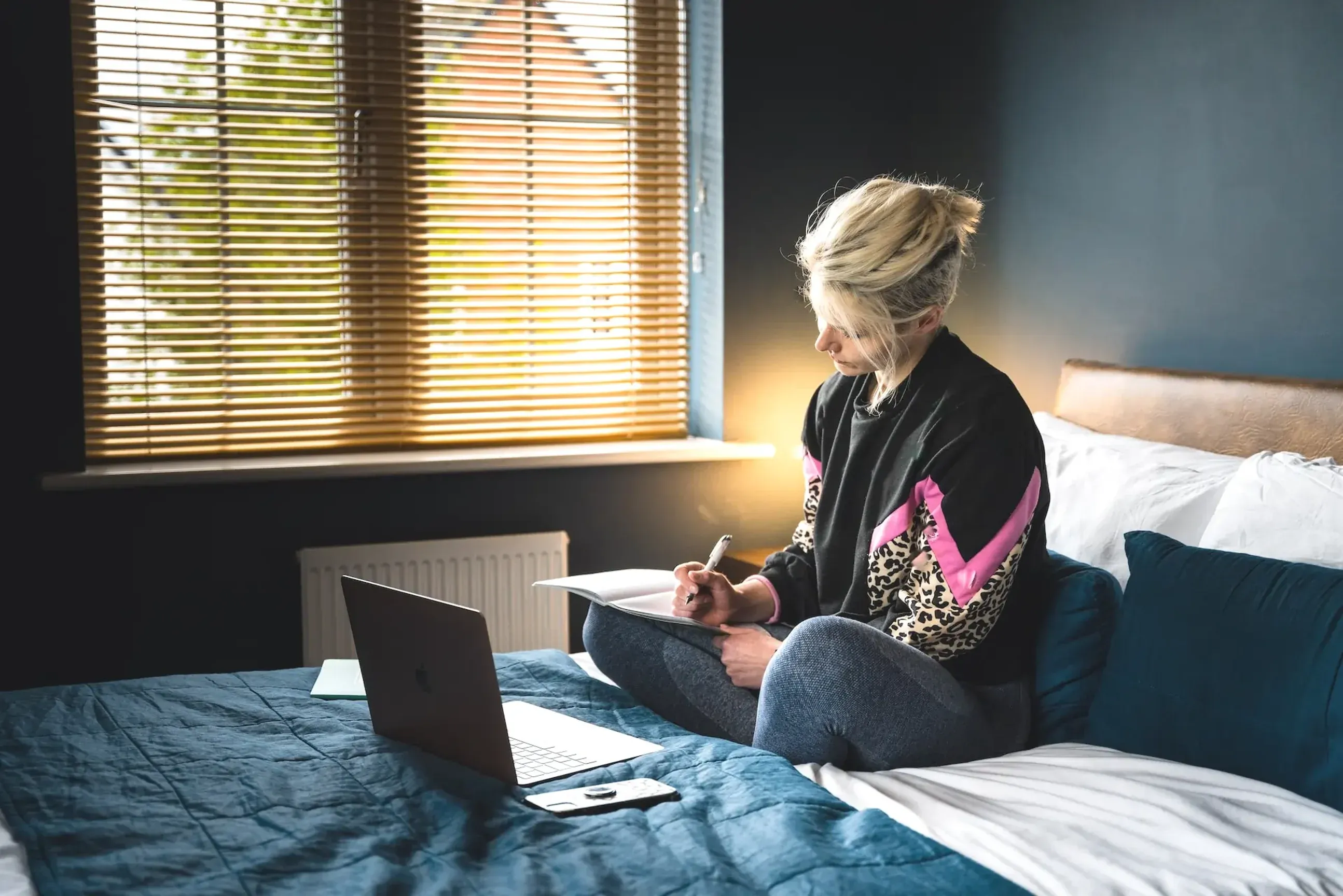 Person sitting on a bed writing in a notebook with a laptop nearby.
