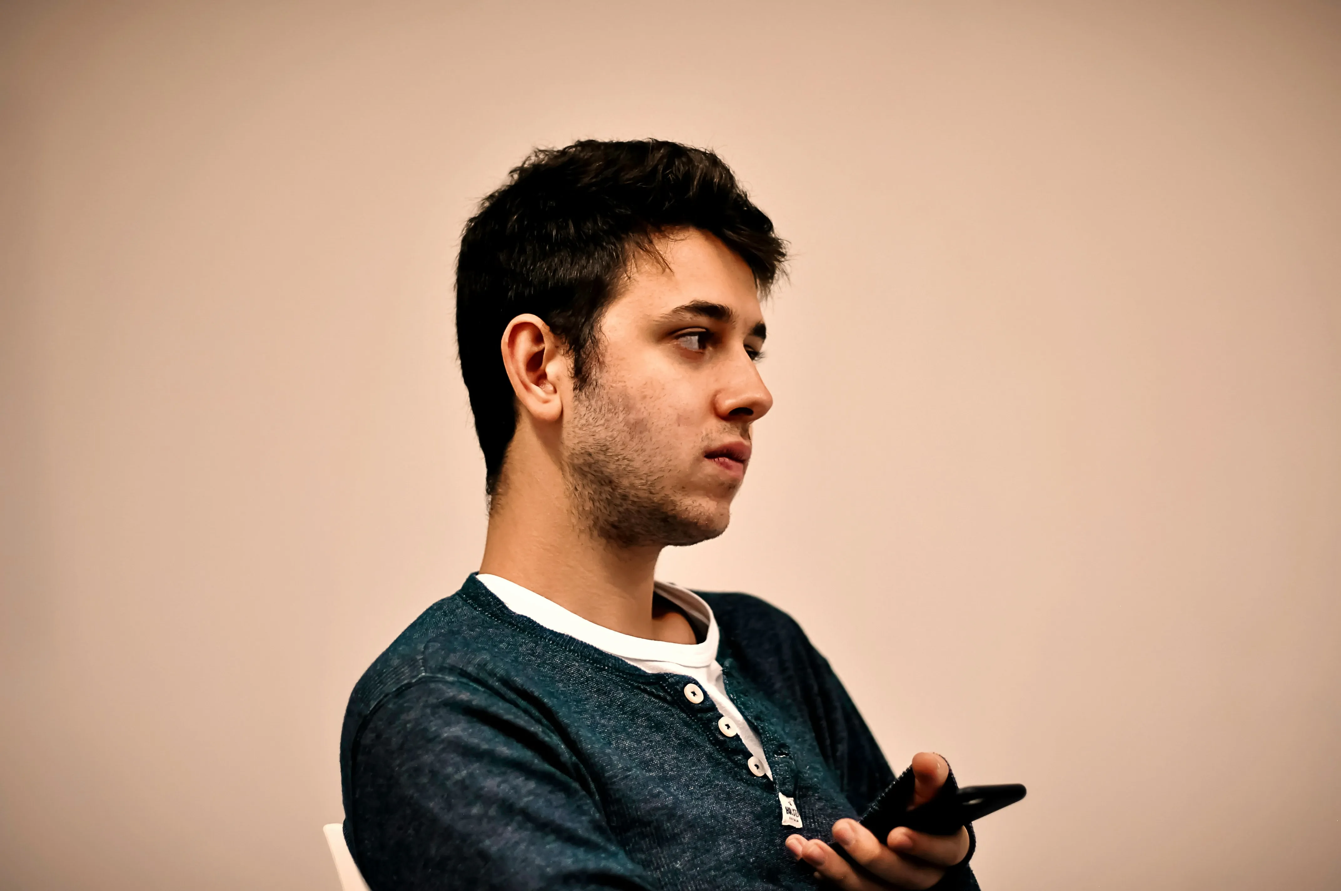 A young man holding a phone and looking off to the side with a thoughtful expression against a neutral background.